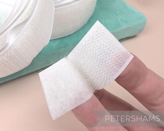 May include: White hook and loop tape, also known as Velcro, is shown in a close-up image. The tape is rolled up and a small piece is being held by fingers to show the texture of the fabric.