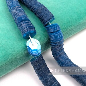 May include: Close-up of a blue sequin necklace with a light blue sequin pendant. The necklace is made of round, flat sequins strung together. The background is a teal green fabric.