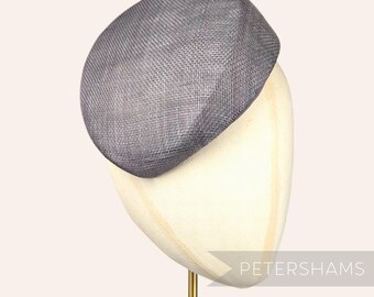 May include: A grey, round, pillbox hat made of woven fabric. The hat is on a white hat stand with a gold metal base. The text "PETERSHAMS" is visible in the lower right corner of the image.