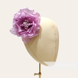 May include: A large, single, purple silk flower with a ruffled edge is displayed on a mannequin head. The flower is a statement piece for a hat or hair accessory.