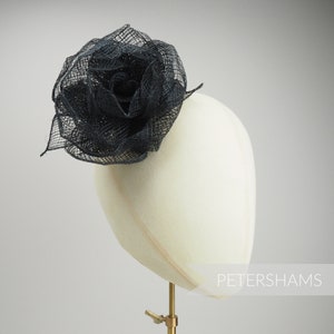 May include: A navy blue fascinator in the shape of a rose, made of a mesh-like material. The headpiece is mounted on a white mannequin head. The word "PETERSHAMS" is printed on the lower right side of the image.