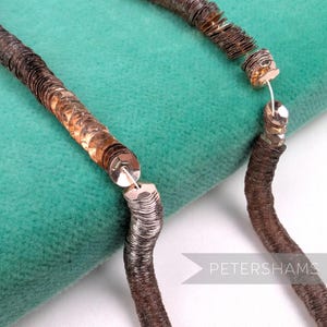 May include: Close-up of a brown sequin necklace with silver-toned clasps. The sequins have a metallic sheen, and the necklace is displayed on a teal-colored surface. The word "PETERSHAMS" is visible in the image.