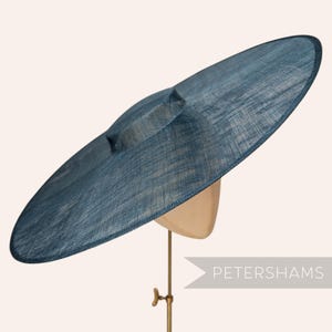 Peut inclure: Un chapeau à large bord d'une teinte bleu profond avec une texture tissée et une calotte plate. Le chapeau est présenté sur un support et porte le texte PETERSHAMS dans le coin inférieur droit. Le design est circulaire et uniforme.