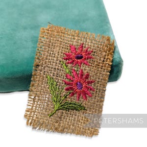 Pode incluir: Um pequeno peda&ccedil;o quadrado de serapilheira com um desenho de bordado floral. O bordado apresenta duas flores cor-de-rosa com centros roxos e folhas verdes. A serapilheira tem uma apar&ecirc;ncia &aacute;spera e texturizada com bordas desfiadas.