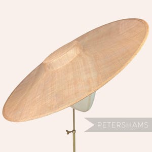 Peut inclure: Un chapeau de soleil beige à larges bords, avec une couronne structurée et un large bord circulaire. Le chapeau est fait de matériau tissé et est présenté sur un support à chapeau. Le texte "PETERSHAMS" est visible.