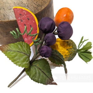 Op de afbeelding: Een handgemaakte fruit broche met een watermeloen, druiven en een ananas. De broche is gemaakt van stof en heeft een groene steel met bladeren.