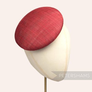May include: A red, round, woven hat on a white hat stand. The hat is tilted to the side. The stand is on a gold metal rod. The text "PETERSHAMS" is visible in the lower right corner of the image.