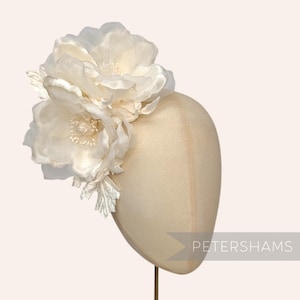 May include: Cream-coloured floral headpiece featuring two large, layered fabric flowers and delicate silver-toned leaves. The headpiece is displayed on a mannequin head. The text "PETERSHAMS" is visible on the right side.