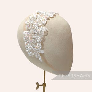 May include: A white floral headband with intricate lace detailing and iridescent accents. The headband is displayed on a mannequin head, showcasing its design and craftsmanship. The text "PETERSHAMS" is visible.