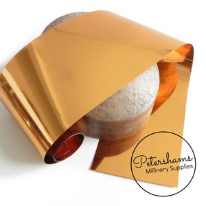 May include: A roll of shiny copper-colored metallic fabric. The fabric is rolled up on a wooden form and is lying on a white surface. Petershams Millinery Supplies is printed on a white circle.