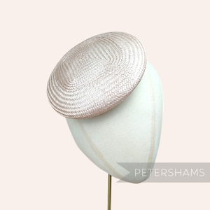 Può includere: Un cappello rotondo intrecciato di colore rosa chiaro su un supporto bianco. Il cappello è un design semplice ed elegante.