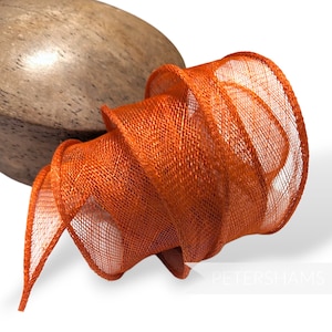 May include: A close-up of a bright orange, woven, fabric ribbon. The ribbon is curled and twisted, creating a decorative effect.