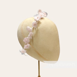 Può includere: Un manichino per cappello giallo pallido è adornato da una fascia floreale. La fascia presenta piccoli fiori rosa chiaro, creando un accessorio elegante. Il manichino è montato su un supporto dorato. I fiori sono un design delicato.