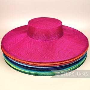 May include: A stack of wide-brimmed hats in various colours, including pink, orange, blue, green, and yellow. The hats are made of a fine, woven material and have a slightly rounded crown. The top hat is a bright pink colour. The text "PETERSHAMS" is visible at the bottom of the image.