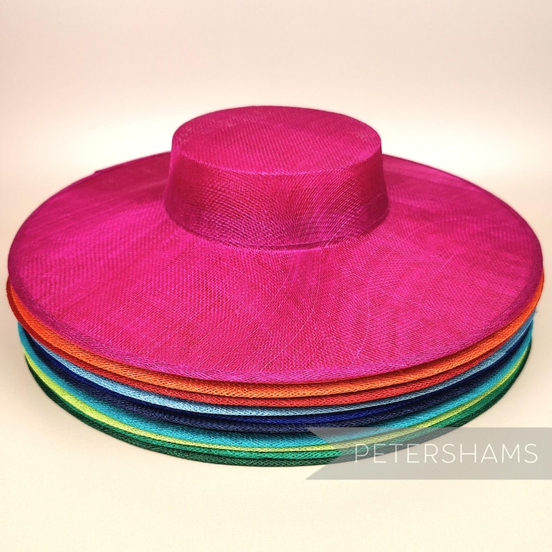 May include: A stack of wide-brimmed hats in various colours, including pink, orange, blue, green, and yellow. The hats are made of a fine, woven material and have a slightly rounded crown. The top hat is a bright pink colour. The text "PETERSHAMS" is visible at the bottom of the image.