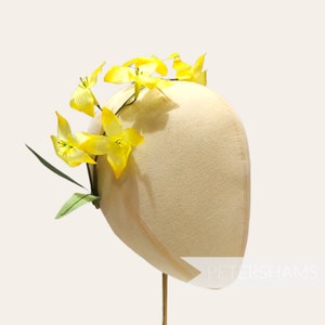 Op de afbeelding: Een gele bloemenkrans met groene bladeren op een beige hoedvorm. De hoed staat op een standaard met de tekst "PETERSHAMS" aan de zijkant.