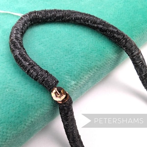 May include: Close-up of a black, curved, sequined trim. The trim is made of small, round, black sequins sewn together. A gold-colored metal clasp connects the trim. The background is a soft, teal-colored fabric. The text "PETERSHAMS" is in the lower right corner.