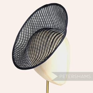 May include: A navy blue, wide-brimmed hat with a woven, grid-like pattern. The hat is displayed on a wooden head form with a gold-colored stand. The text "PETERSHAMS" is visible on the base.