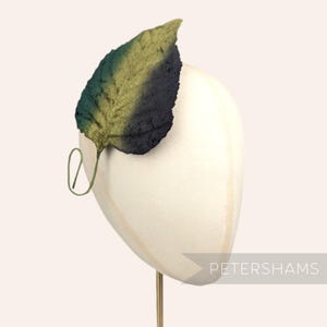 Könnte beinhalten: Ein grünes, goldenes und schwarzes Blatt-Haarschmuck. Das Blatt ist aus Stoff und hat eine strukturierte Oberfläche. Der Schmuck befindet sich auf einem weißen Mannequin-Kopf.