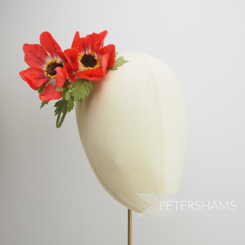 May include: A red flower hair clip with two red flowers and green leaves. The clip is on a white mannequin head.