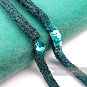 May include: Close-up of a teal sequin trim. The trim is made of small, round sequins sewn onto a fabric backing. The sequins are a mix of teal and dark teal, creating a shimmering effect. The trim is displayed against a teal background.