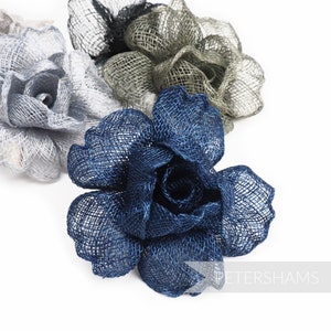 May include: Close-up of three fabric flower embellishments in shades of blue, gray, and silver. The flowers are made of a woven material, with a rose-like design. The flowers are on a white background.