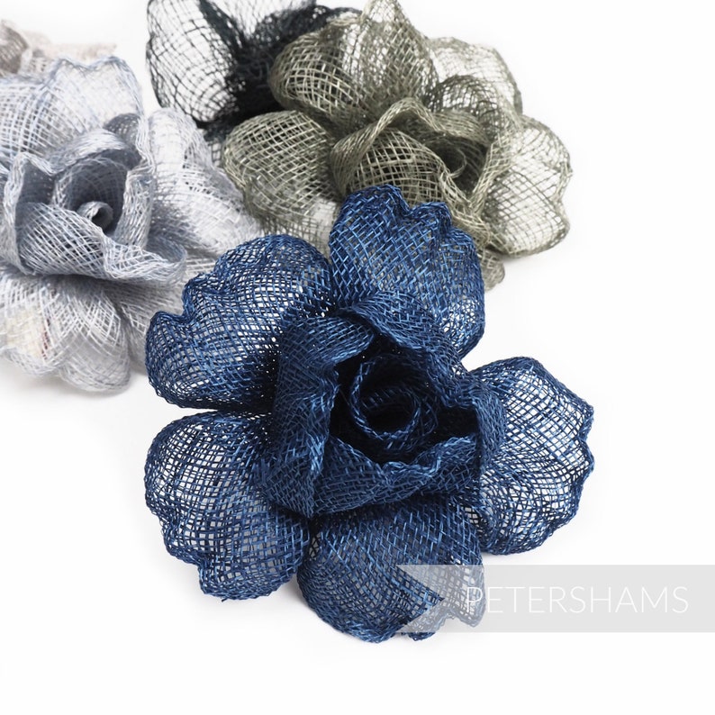 May include: Close-up of three fabric flower embellishments in shades of blue, gray, and silver. The flowers are made of a woven material, with a rose-like design. The flowers are on a white background.