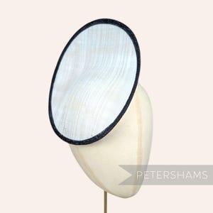 May include: A white, circular hat with a black trim, displayed on a mannequin head. The hat has a woven texture and is mounted on a gold-colored stand. The text "PETERSHAMS" is visible on the right side of the image.