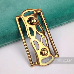 Könnte beinhalten: Goldfarbene Metallschnalle mit offenem Design. Die Schnalle hat eine rechteckige Form mit abgerundeten Kanten und zwei kreisförmigen Verzierungen. Das Design umfasst ovale und rautenförmige Aussparungen. Für einen Gürtel oder ein Accessoire.