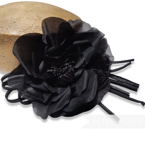 May include: A black fabric flower with layered petals and a textured center. The flower is attached to a base with several long, thin, black fabric strips. The flower is set against a light-colored, wooden form.
