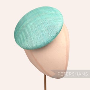 May include: A teal blue round hat made of woven straw, displayed on a hat stand. The hat is a simple, elegant design.