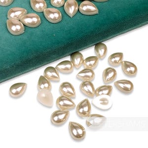 May include: A large quantity of off-white pearlescent teardrop-shaped beads are scattered on a white surface. Some of the beads are on a teal velvet surface.