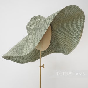 May include: A large, wide-brimmed, green straw hat with a woven texture. The hat is displayed on a gold metal stand.