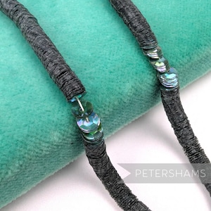 May include: Close-up of a dark gray sequin trim with iridescent sequins. The trim is a thick, cord-like material with a textured surface. The iridescent sequins are round and reflect multiple colors. The trim is on a teal background.