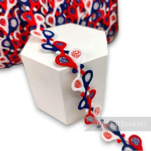 May include: Red, white, and blue embroidered trim featuring a lacrosse stick design. The trim is shown wrapped around a white hexagonal display stand. The text "PETERSHAMS" is visible in the lower right corner of the image.