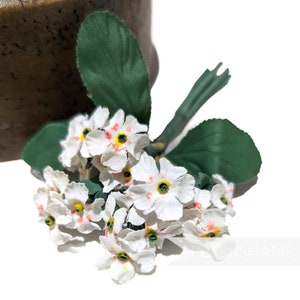 Può includere: Un mazzo di fiori bianchi in tessuto con centri arancioni e foglie verdi. I fiori sono disposti a grappolo e hanno un aspetto delicato e vintage.