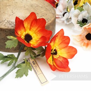 May include: Two orange and yellow artificial flowers with green stems and leaves. The flowers have a yellow center with a dark brown center. A brown tag with the text "NO. 545 MFD. Made in German Democratic Republic" is visible in the image.