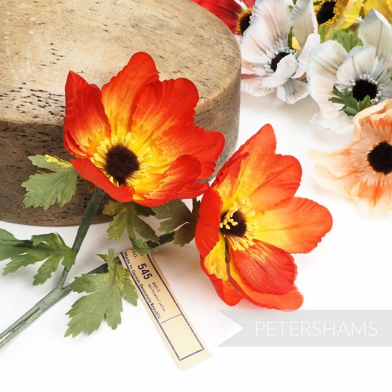 May include: Two orange and yellow artificial flowers with green stems and leaves. The flowers have a yellow center with a dark brown center. A brown tag with the text "NO. 545 MFD. Made in German Democratic Republic" is visible in the image.