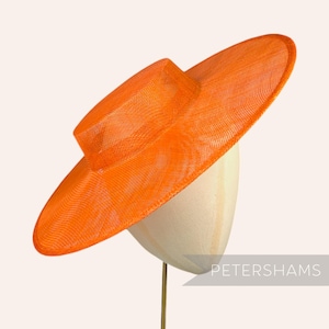 Könnte beinhalten: Ein großer, leuchtend orangefarbener Hut mit breiter Krempe aus geflochtenem Stroh. Der Hut sitzt auf einer Hutform.