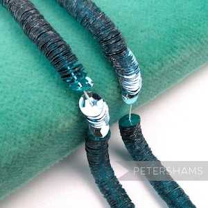 May include: Close-up of two strands of teal and silver sequins, connected by a small, clear, circular clasp. The sequins are arranged in a spiral pattern, creating a textured effect. The background is a soft, teal fabric.