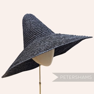 Könnte beinhalten: Ein dunkelmarineblauer Sonnenhut mit breiter Krempe und hohem, konischem Kopf. Der Hut ist aus gewebtem Material gefertigt und wird auf einem Schaufensterkopf präsentiert. Der Text "PETERSHAMS" ist sichtbar.