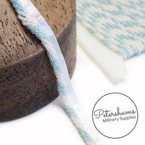 May include: Close-up of white ribbon with a beaded edge in alternating white and light blue. The ribbon is on a wooden surface. Additional fabric samples in white and light blue are in the background. The Petershams Millinery Supplies logo is in the lower right corner.