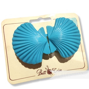以下が含まれることがあります： 「Butt Trim Petershams」と書かれたカードに、ターコイズブルーのプラスチック製の貝殻型のヘアクリップが2つ付いている。