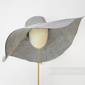 May include: A wide-brimmed sun hat with black and white stripes, displayed on a mannequin head. The hat has a large, floppy brim and a classic striped pattern. The hat is supported by a gold-colored stand.