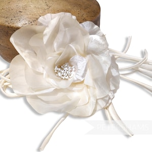 May include: Ivory fabric flower with layered petals and a cluster of white beads at the center. The flower is attached to a bundle of thin, cream-colored ribbons. A wooden form is visible in the background.