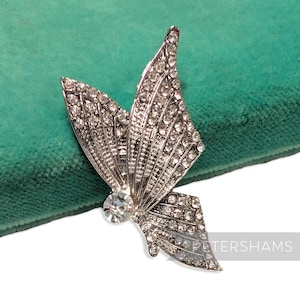 May include: A silver-toned butterfly brooch with clear crystal accents. The brooch features a large central crystal and smaller crystals outlining the wings. The wings have a detailed, textured design. The brooch is set against a green background.