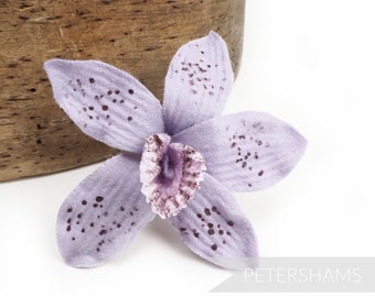 May include: A light purple orchid flower with dark purple speckles. The flower is made of fabric and has a velvety texture.