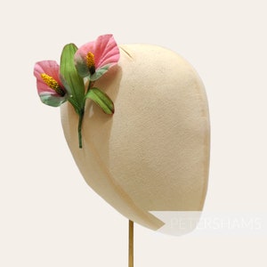 May include: A cream-colored hat with a decorative arrangement of pink and green artificial flowers. The flowers have yellow stamens and are attached to the side of the hat. The hat is displayed on a gold-colored stand.