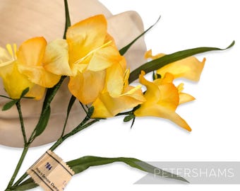 May include: A cluster of bright yellow artificial flowers with green stems and leaves. The flowers are in front of a beige hat. A tag with the text "Made in German Democ" is attached to the stem. The word "PETERSHAMS" is in the bottom right corner.