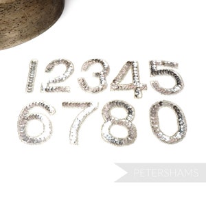 May include: Ten silver sequined iron-on patches in the shape of numbers 0 through 9. The patches are on a white background.